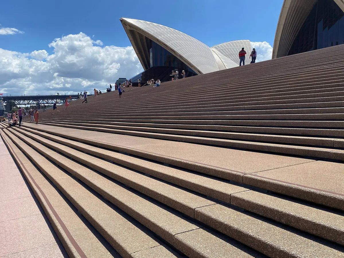 The broad granite steps of the Sydney Opera House rising towards the shells, with visitors sitting on them under a blue sky